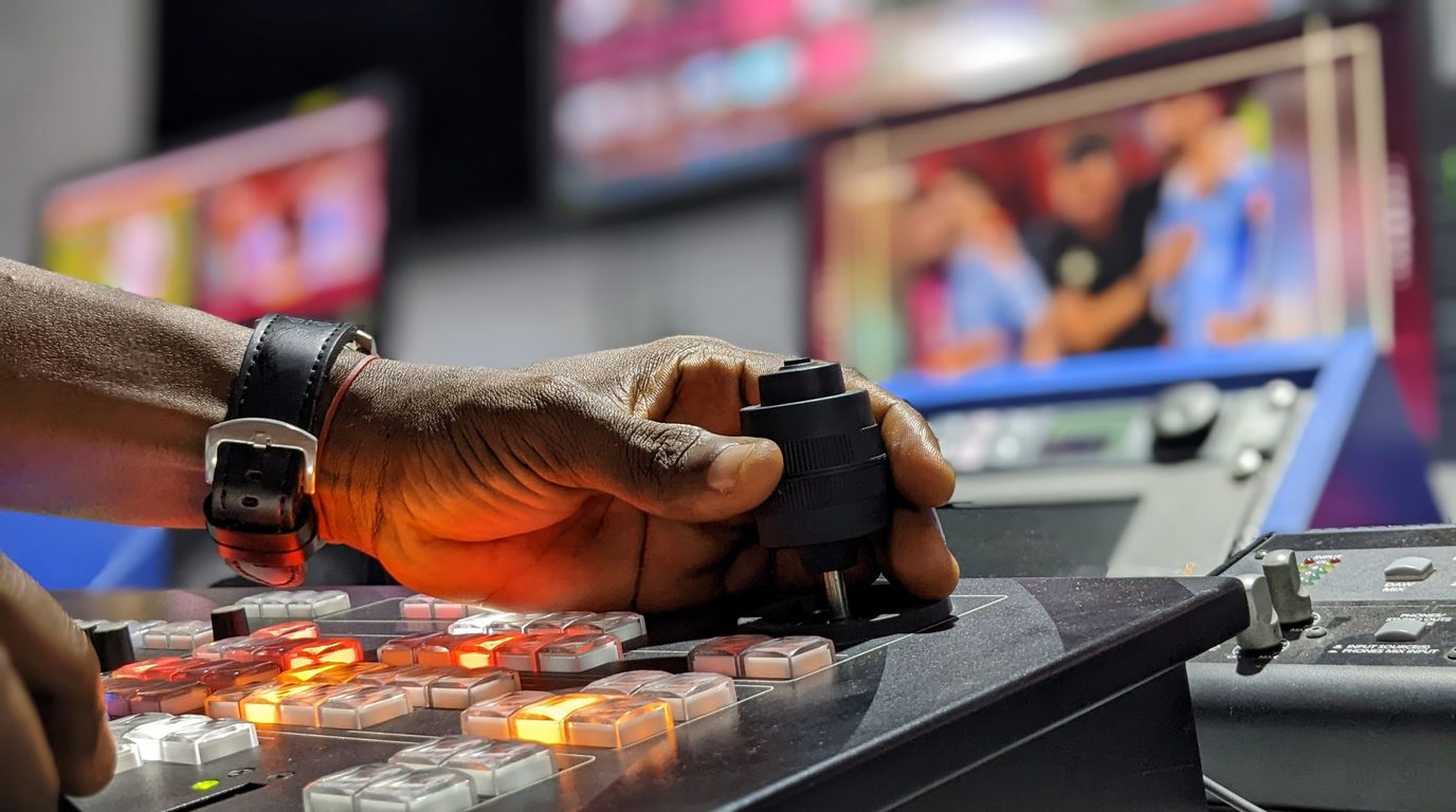 Stock photo: an operator's hand on a video switcher, with live monitors in the background